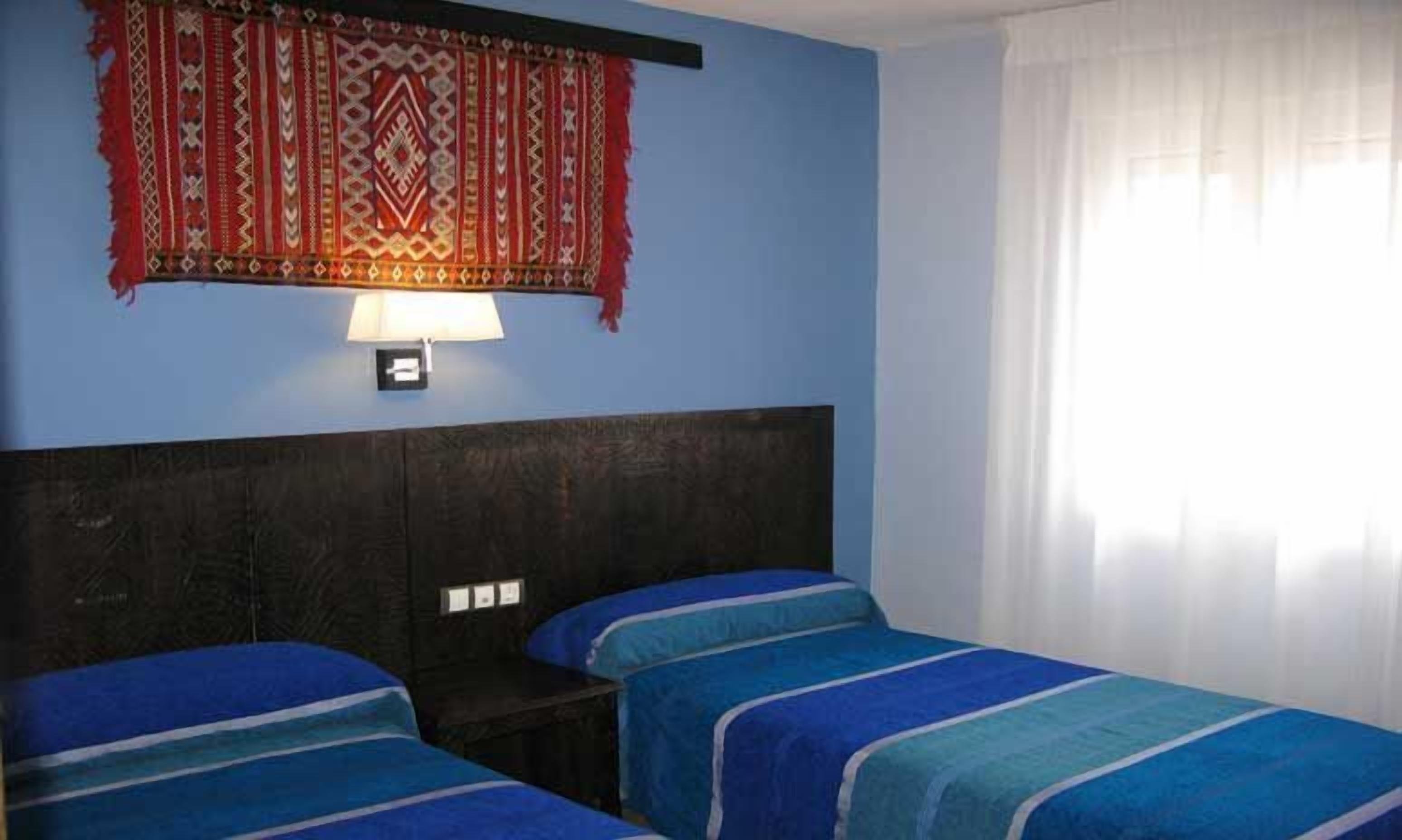 A room with two beds, a dark headboard, a wall-mounted lamp, and a window with curtains.