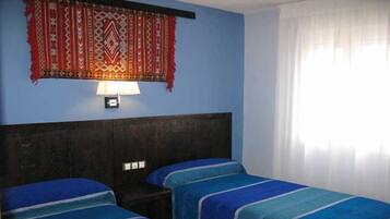 A room with two beds, a dark headboard, a wall-mounted lamp, and a window with curtains.