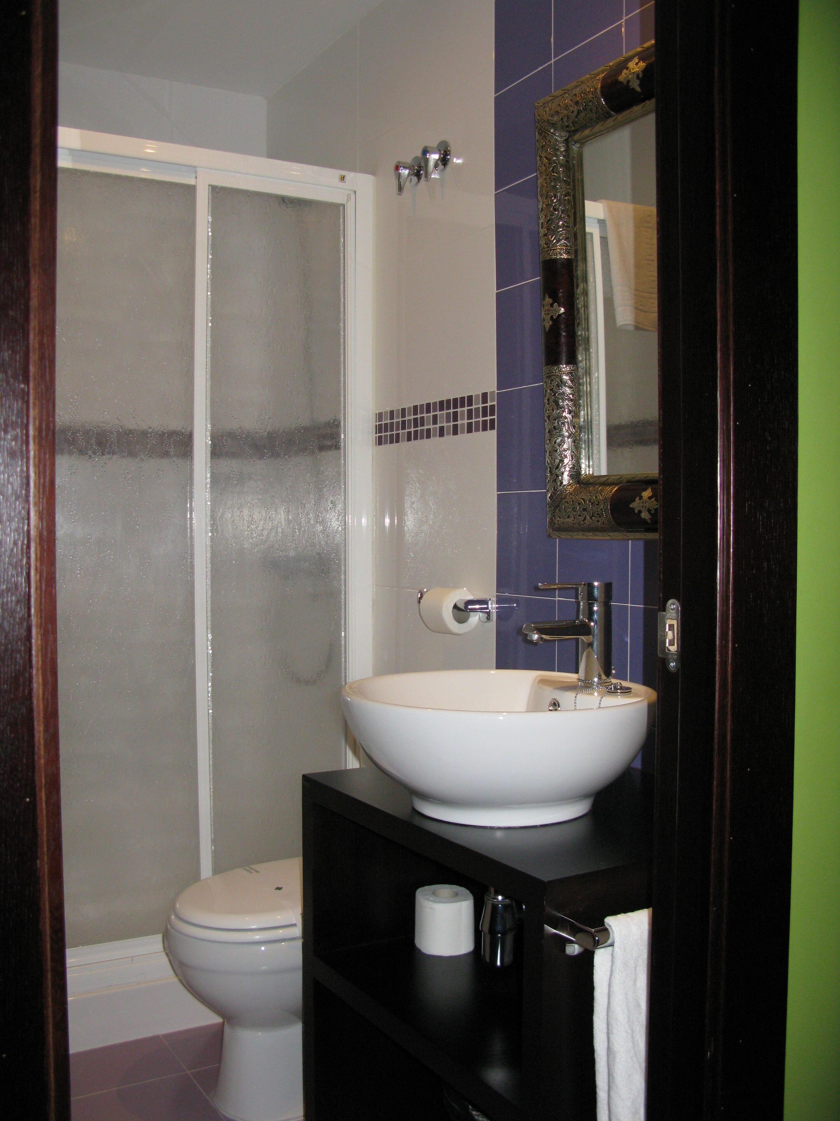 Casa de banho com uma sanita, uma lavatório branco, chuveiro com portas de vidro fosco e um espelho com moldura ornamentada.