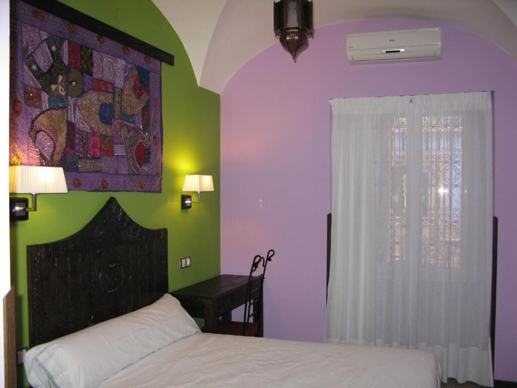 Un dormitorio con cama, dos lámparas de noche, aire acondicionado de pared y ventana con cortinas traslúcidas.