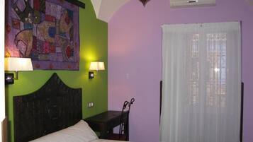 Un dormitorio con cama, dos lámparas de noche, aire acondicionado de pared y ventana con cortinas traslúcidas.