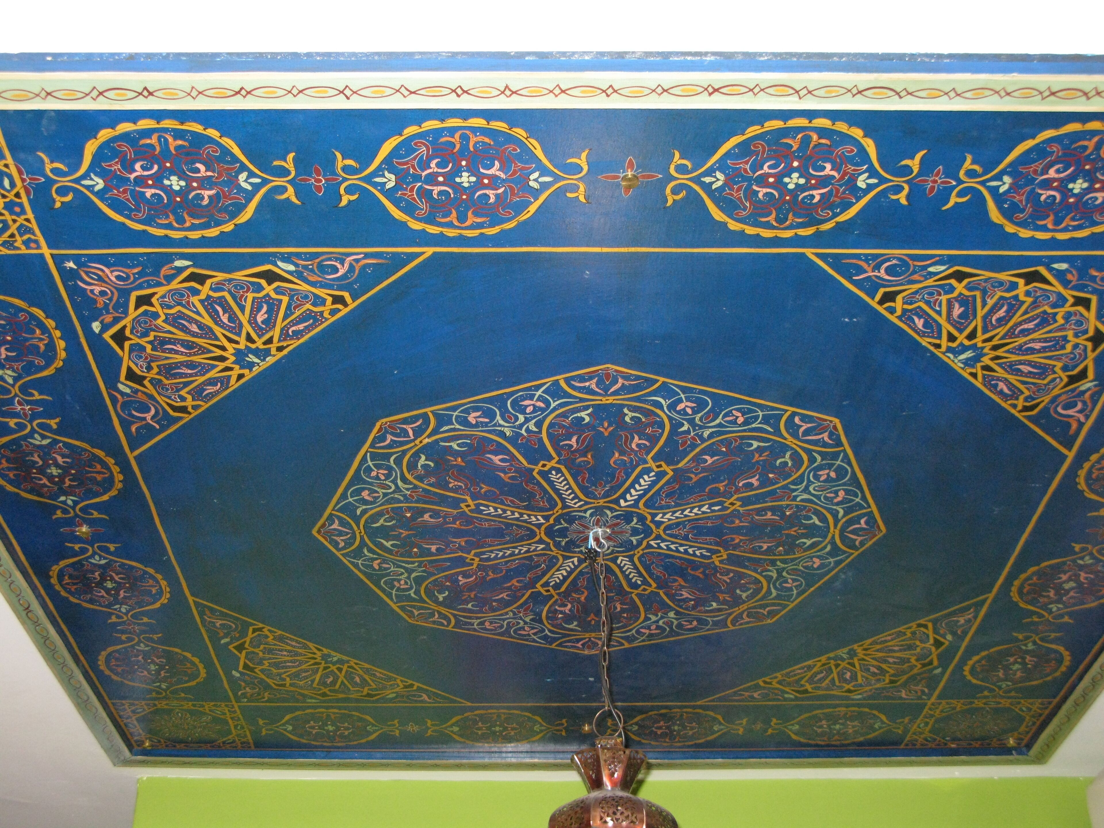 A ceiling with intricate geometric and floral patterns, a central hanging light fixture, and a green wall.