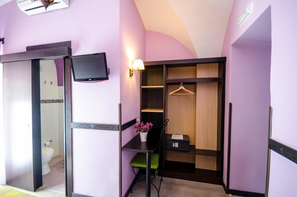 A compact room with a desk, a chair, a small table with a potted plant, a hanging coat rack, and a wardrobe with shelves.