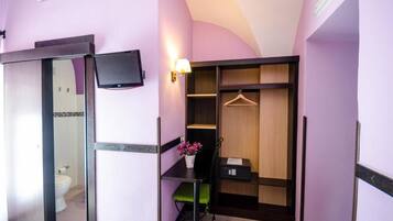 A compact room with a desk, a chair, a small table with a potted plant, a hanging coat rack, and a wardrobe with shelves.