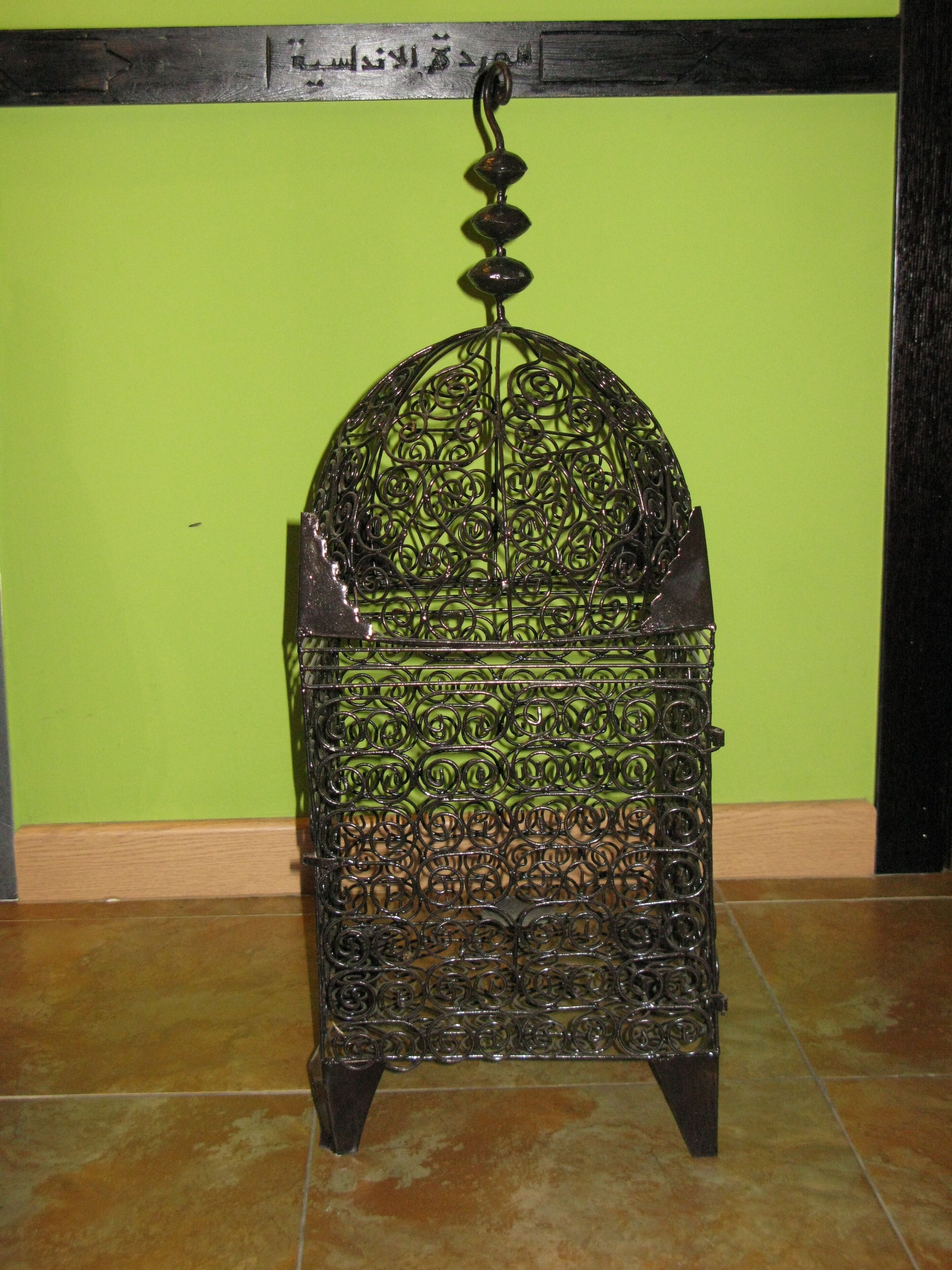 Un mueble metálico con diseños intrincados, un gancho en la parte superior y una pared verde al fondo.