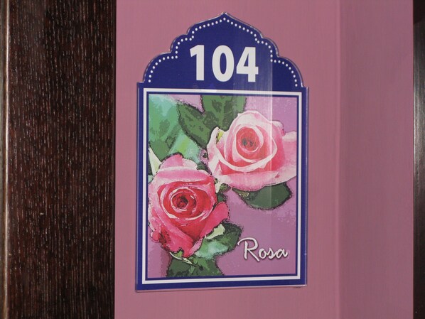 Sininen kyltti, jossa on numero 104, on kiinnitetty seinään. Kyltin alla on kaksi vaaleanpunaista ruusua ja niiden alapuolella sana »Rosa».