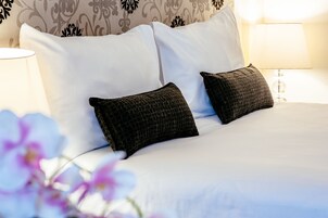 A neatly made bed with white linens and two dark pillows, a bedside lamp, and a floral arrangement.
