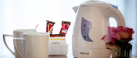 A white electric kettle with a capacity indicator, two white mugs, a box of tea bags, and a pink rose in a glass vase on a countertop.