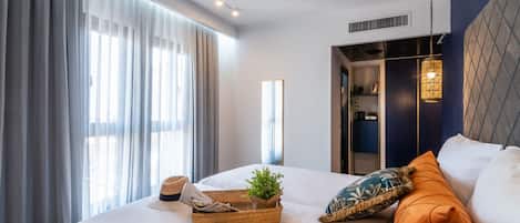 A modern hotel room with a large bed, a side table with a plant and a hat, and a view of a hallway with a mirror and a wardrobe.