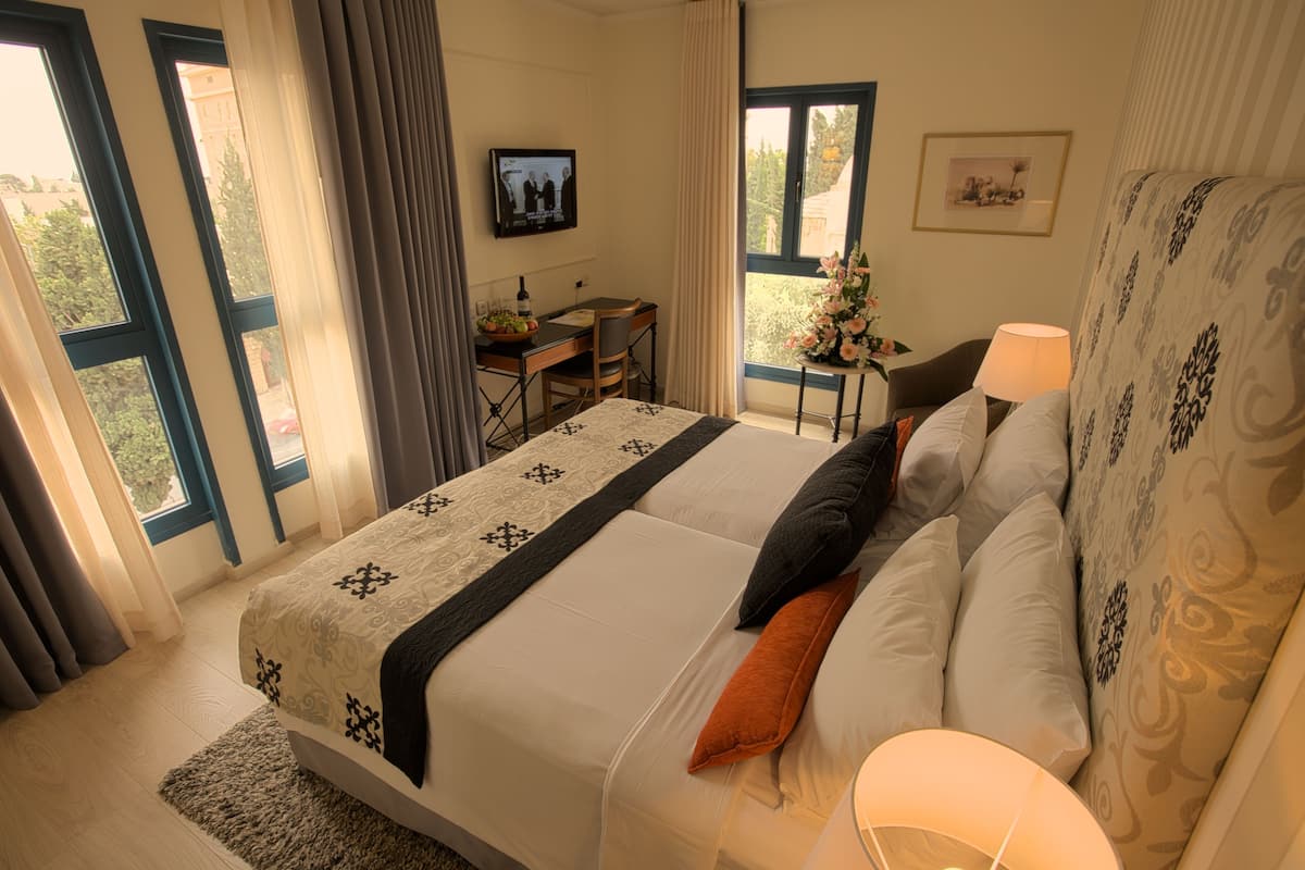 A hotel room with a large bed, a desk, a chair, a TV, and a window with a view of greenery.