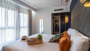 A modern hotel room with a large bed, a bedside table with a plant, a basket, and a hat. There is a window with sheer curtains and a view of the outside. - The Eldan Hotel (Jerusalem)