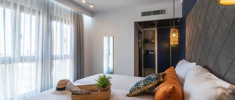 Una habitación de hotel moderna con una cama grande, una mesita de noche con una planta, una cesta y un sombrero. Hay una ventana con cortinas translúcidas y vistas al exterior.