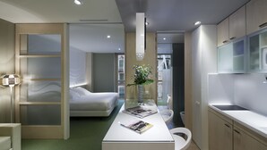 Una habitación de hotel moderna con una cama, un escritorio con un jarrón de flores, una silla blanca y una zona de cocina con armarios y un lavamanos.