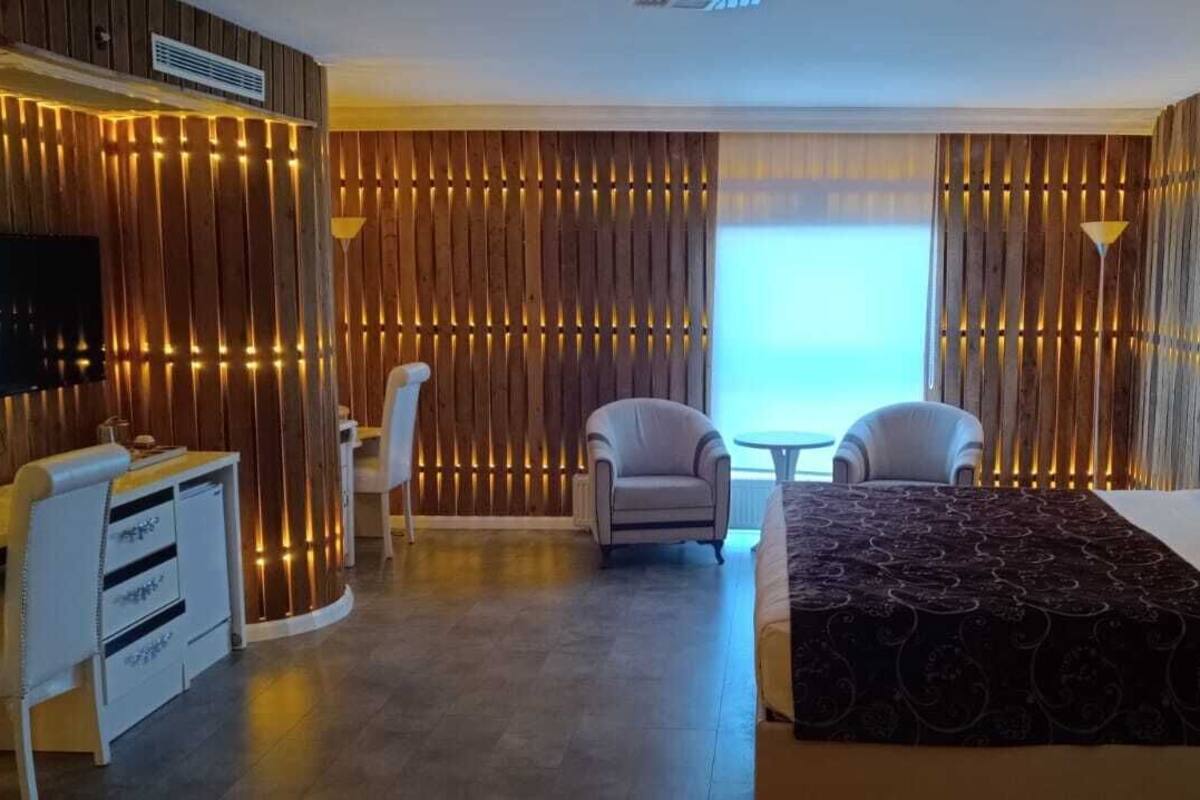 A modern hotel room with a wooden panel wall, a flat-screen TV, a desk with drawers, two armchairs, a sofa, and a large bed with a patterned bedspread.