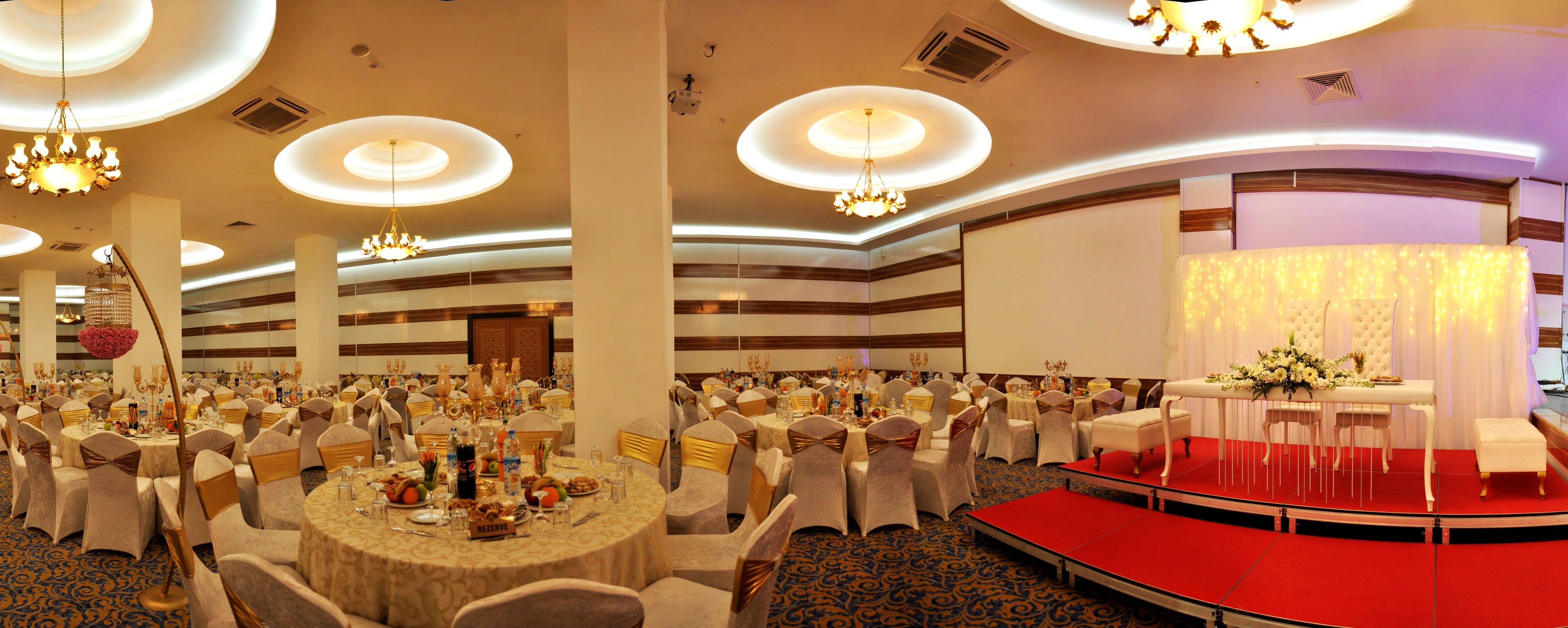 Un salón de banquetes con mesas redondas, sillas, un escenario y candelabros.