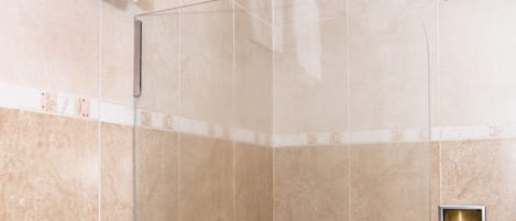 Ducha con cerramiento de vidrio, cabezal de ducha de mano, toallero y un pequeño panel de ventilación en la pared.