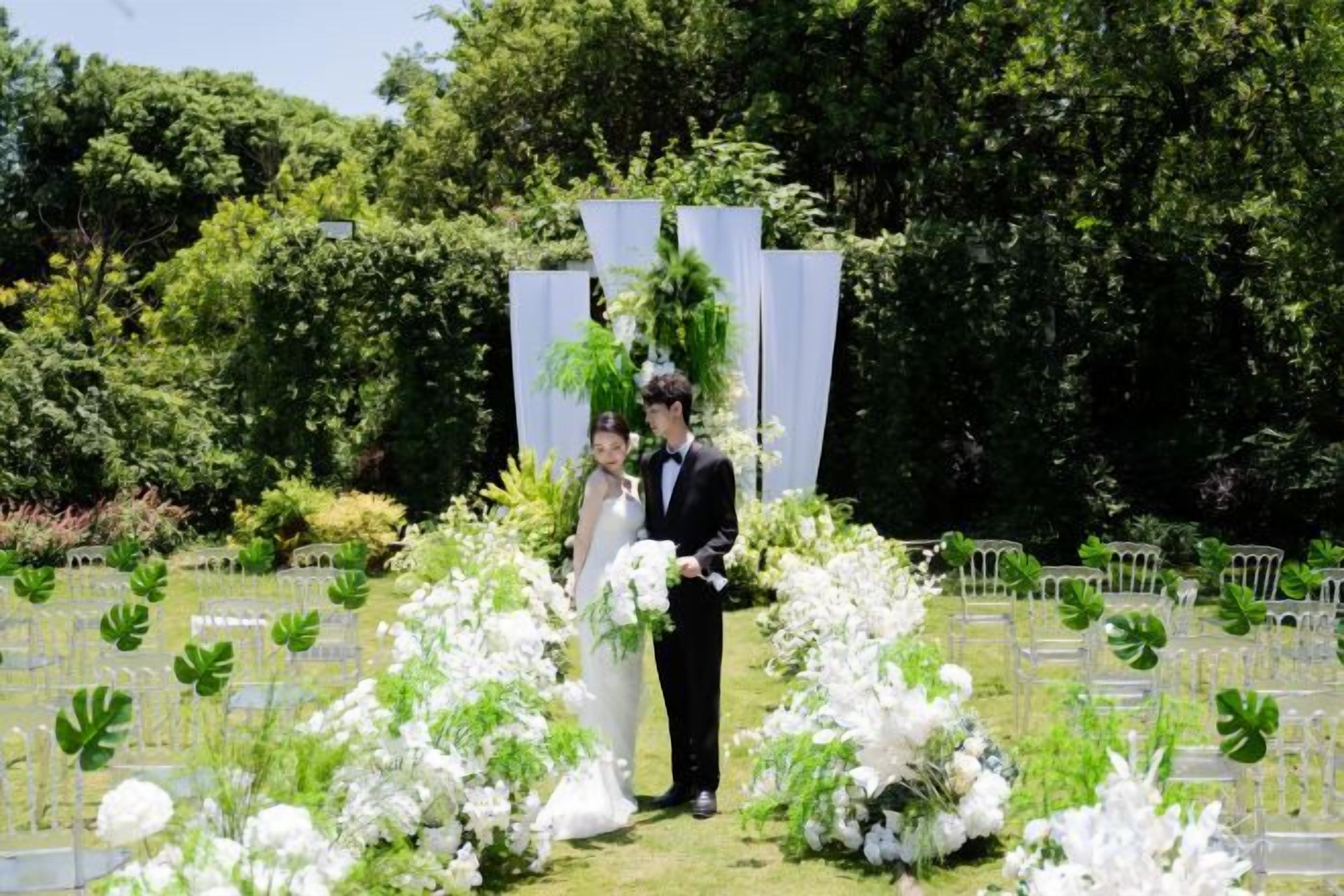 Un couple se tient dans un jardin, avec des compositions florales blanches et une clôture blanche en arrière-plan.