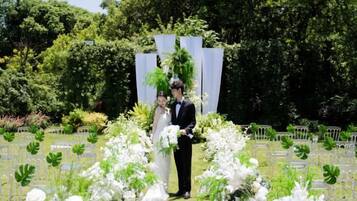 Un couple se tient dans un jardin, avec des compositions florales blanches et une clôture blanche en arrière-plan.