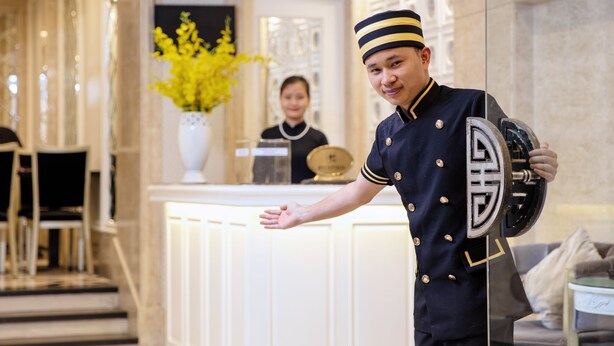 A hotel reception area with a reception desk, a vase with yellow flowers, and a person in uniform holding a camera. - Classy Boutique Hotel (Hanoi)