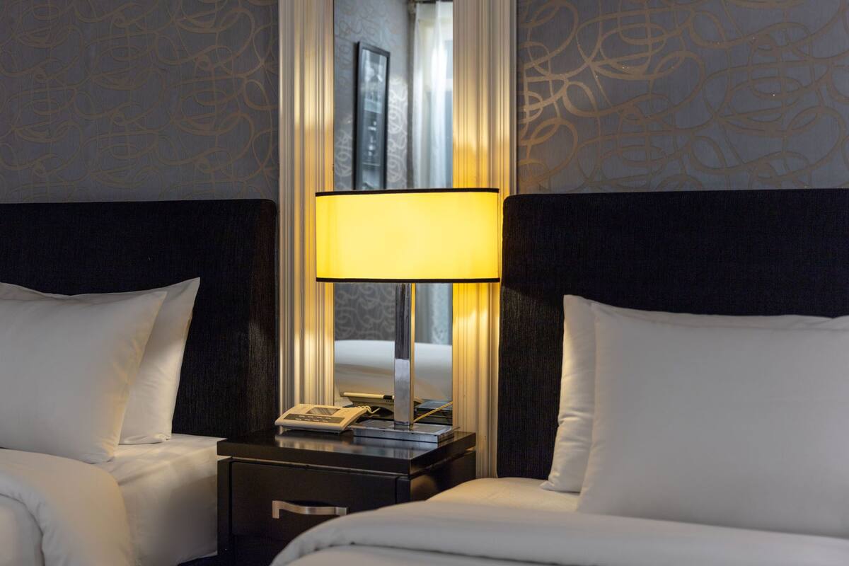 Habitació d'hotel amb dos llits, una tauleta de nit amb una làmpada i un mirall que reflecteix l'espai.