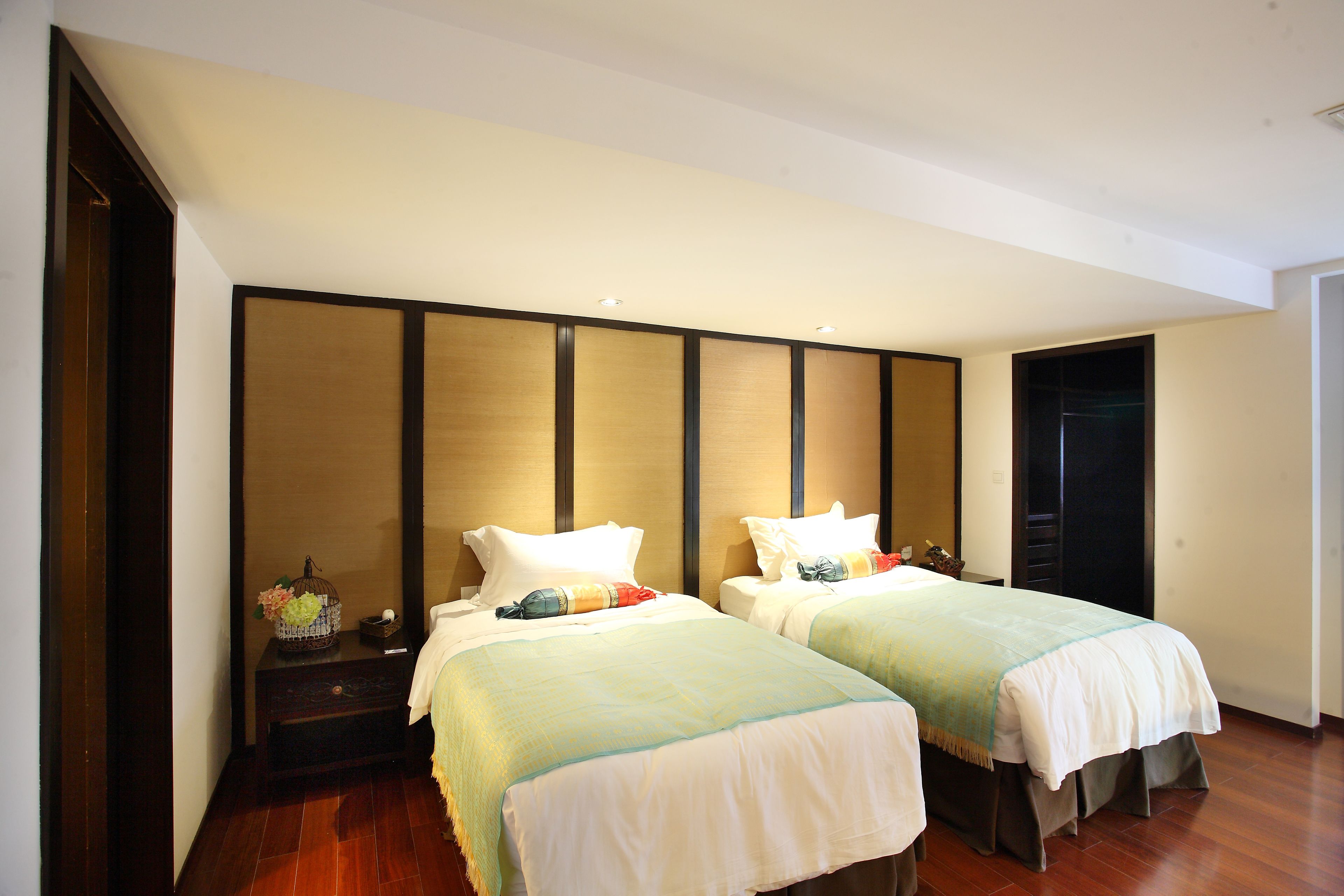 Habitación de hotel con dos camas, cabeceros de madera, una mesita de noche con lámpara y una pequeña mesa con un objeto decorativo.