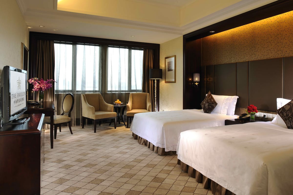 A hotel room with two beds, a desk with a TV, a chair, a small table with a vase of flowers, and a large window with curtains.