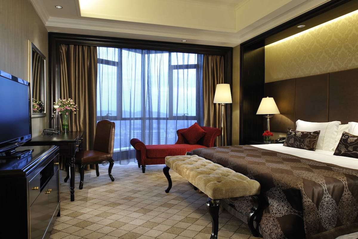 A hotel room with a large bed, a desk with a television, a chair, and a view of the city through large windows.