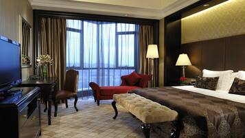 A hotel room with a large bed, a desk with a television, a chair, and a view of the city through large windows.