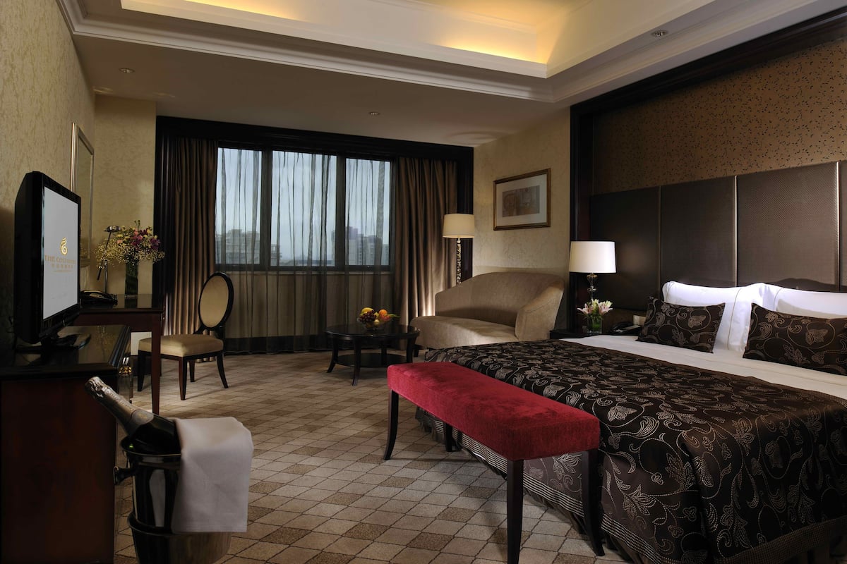 A hotel room with a bed, a desk, a chair, a small table with a vase of flowers, and a window with curtains.