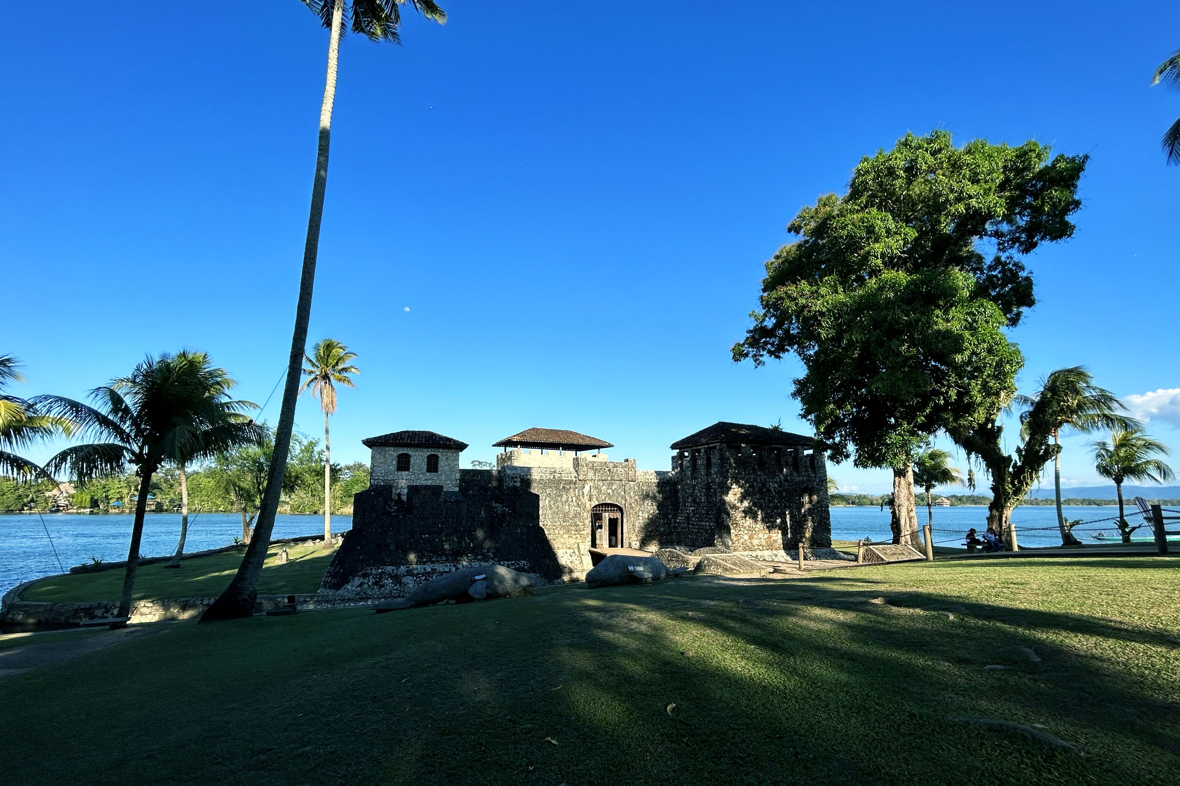 Un edificio histórico de piedra con una torre central y dos estructuras más pequeñas, rodeado de palmeras y una masa de agua.