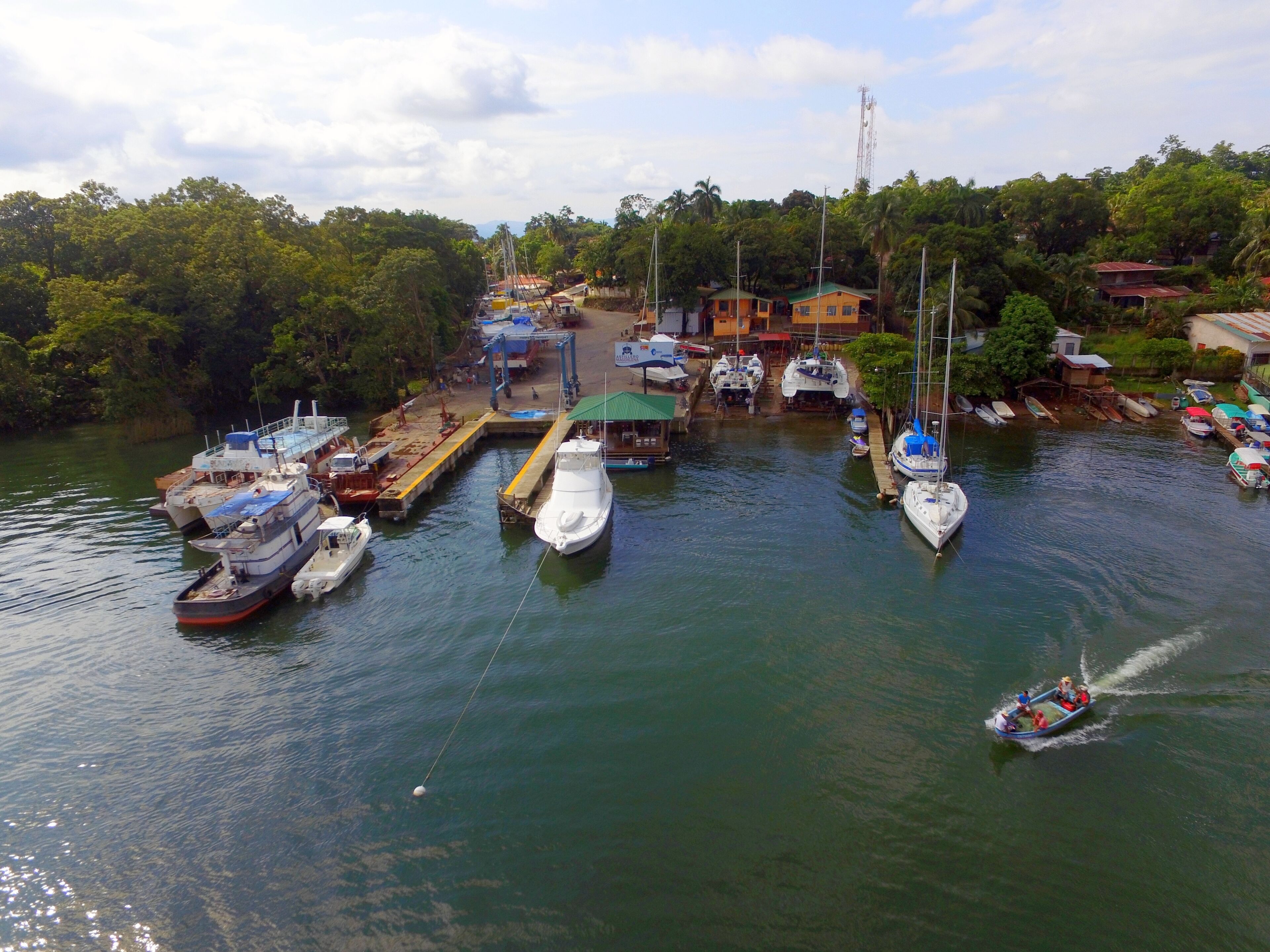 Un port de plaisance où sont amarrés différents types de bateaux, une petite embarcation qui se déplace sur l’eau, et des bâtiments en arrière-plan.