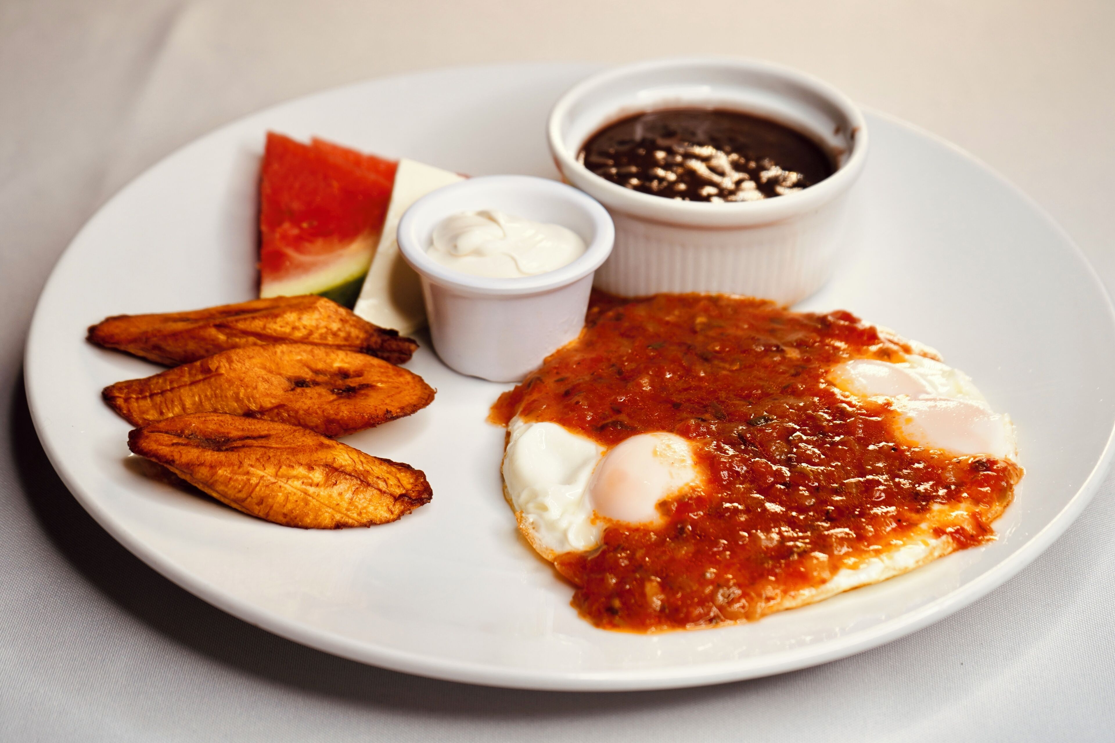 A plate with a fried egg, plantains, a slice of watermelon, and two small bowls of sauce.