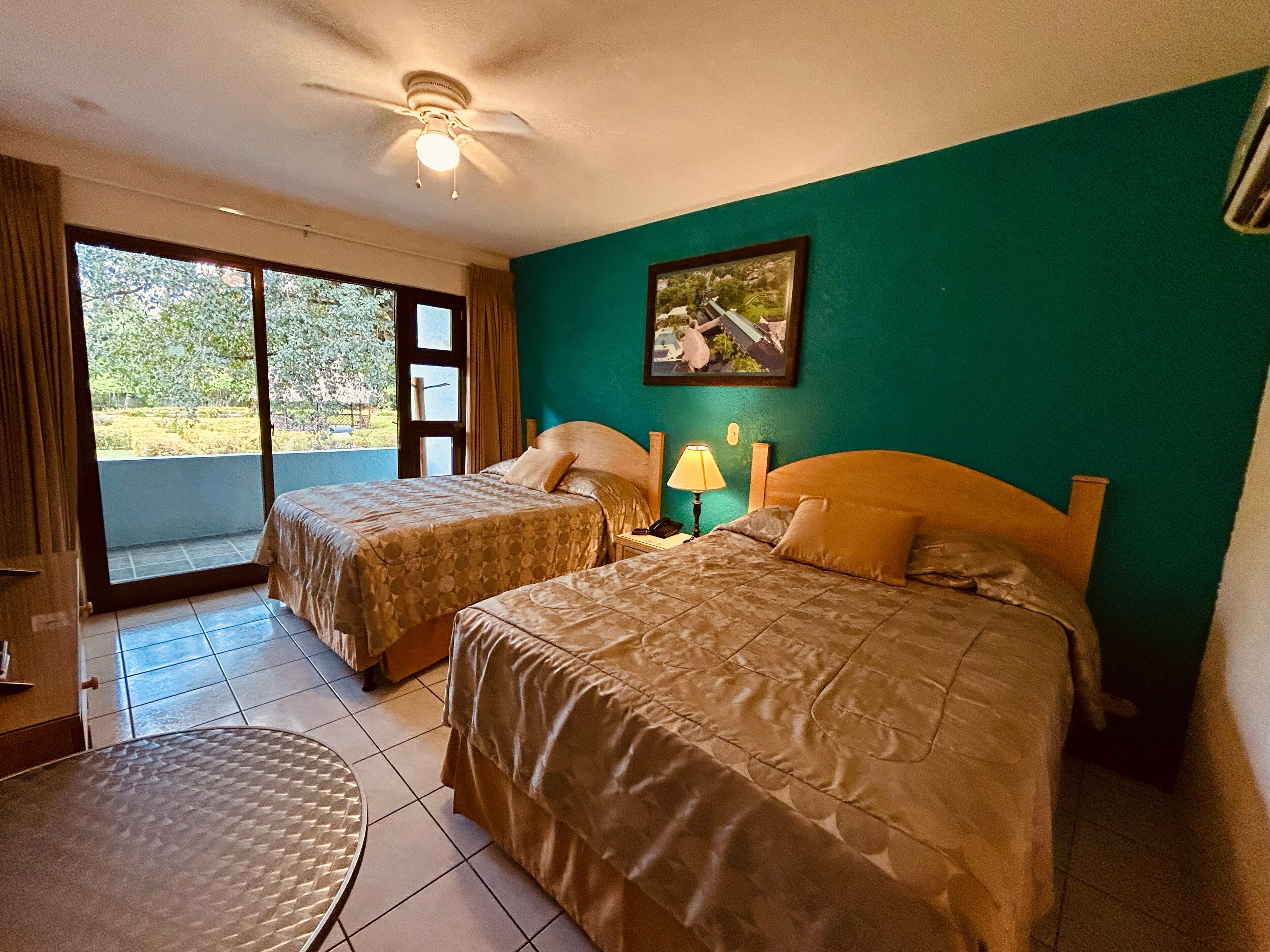 Camera d'albergo con due letti, una ventola a soffitto, una finestra vista verde e un tavolo.
