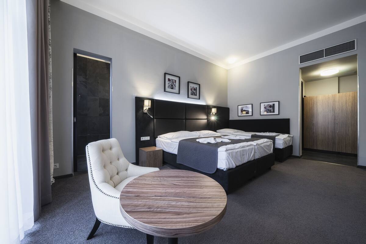 A modern hotel room with a large bed, a round wooden table, a white armchair, and a dark headboard.