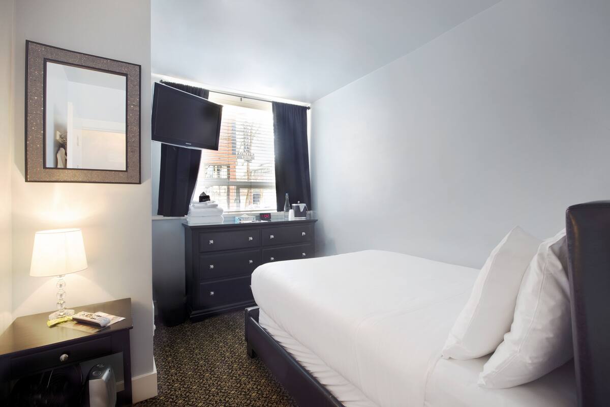 A hotel room with a bed, a nightstand with a lamp, a dresser, a mirror, and a window with curtains.