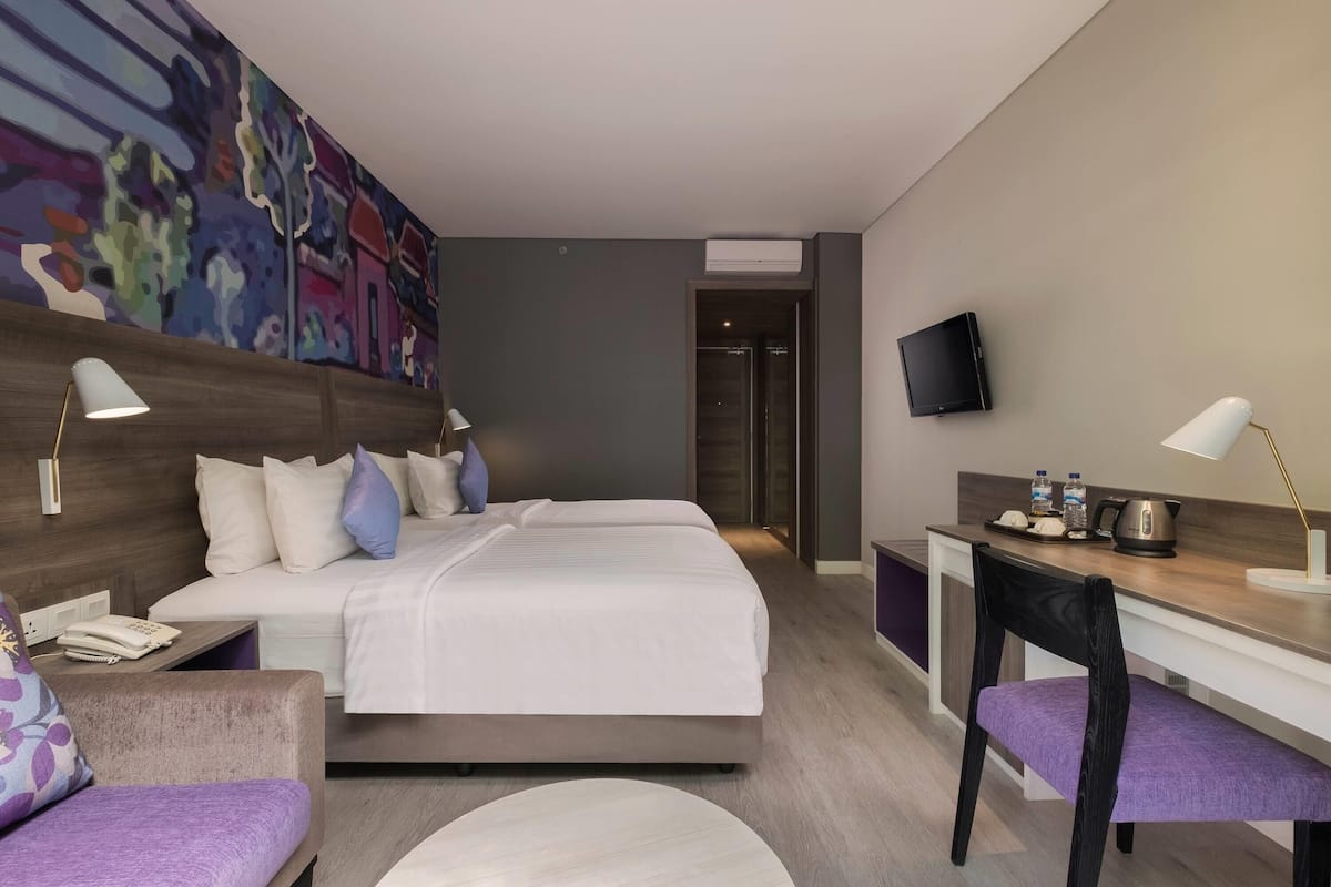 A modern hotel room with a large bed, a desk, a chair, a TV, and a colorful abstract painting on the wall.