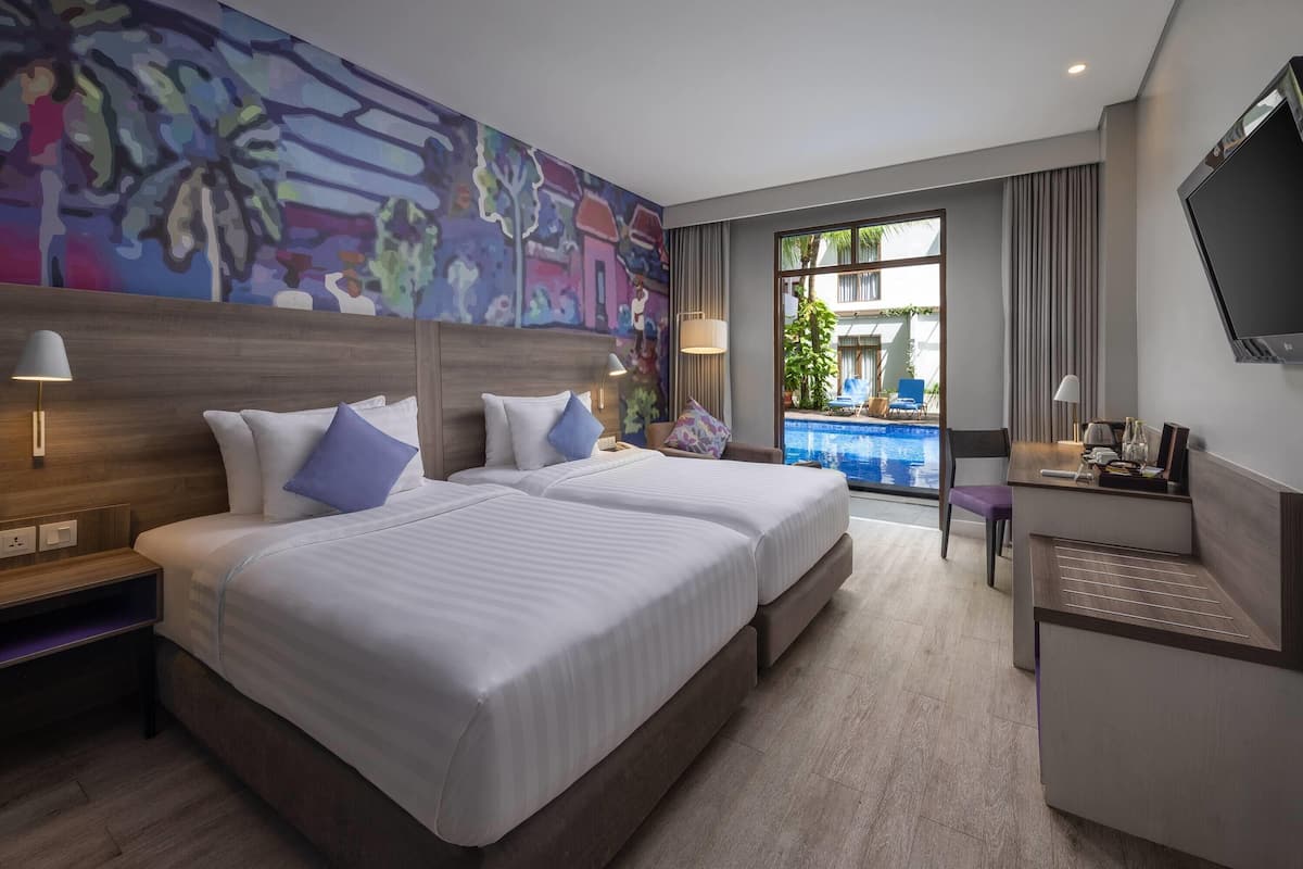 A hotel room with two beds, a desk, a TV, and a large window with a view of a pool and buildings.