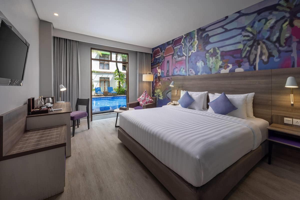 A modern hotel room with a large bed, a desk, a chair, a TV, and a view of an outdoor swimming pool.