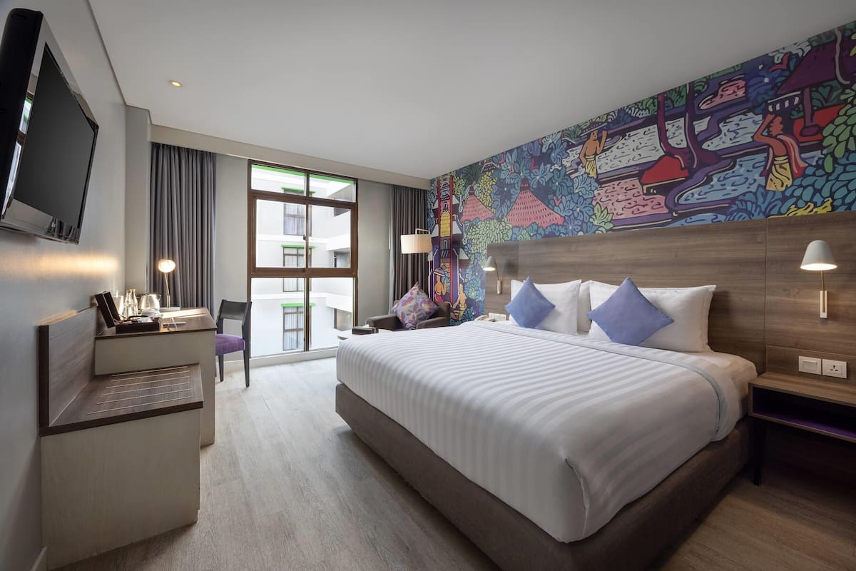 A hotel room with a bed, desk, chair, TV, and a colorful mural on the wall.