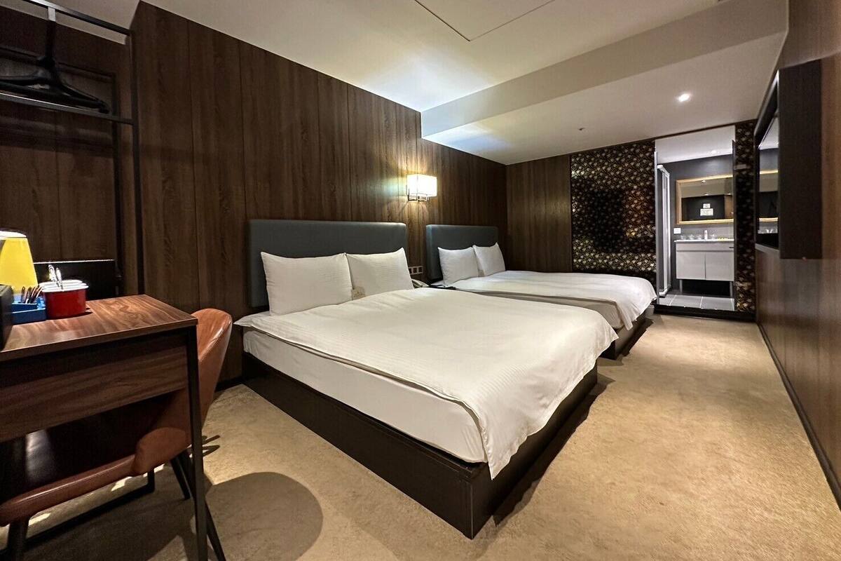 A hotel room with two beds, a desk, a chair, a lamp, and a bathroom visible through an open door.