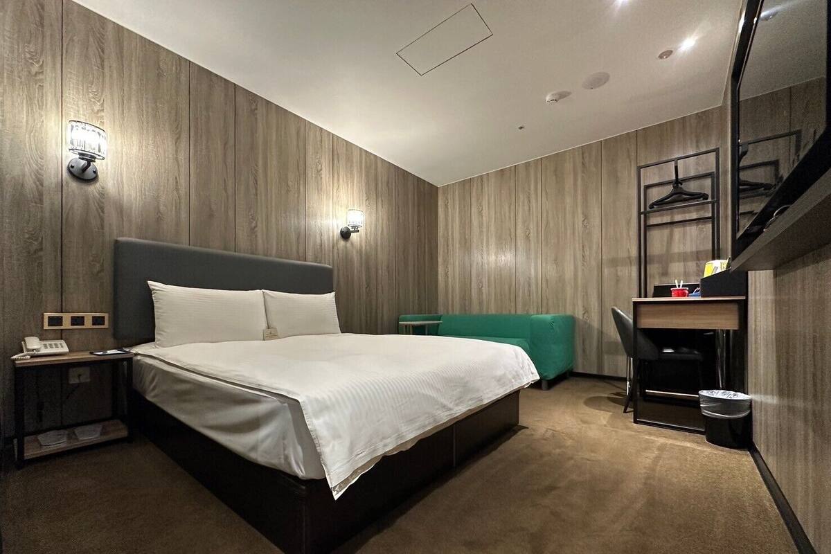 A modern hotel room with a large bed, a desk, and a chair. There is a telephone on the nightstand and a trash can near the desk.