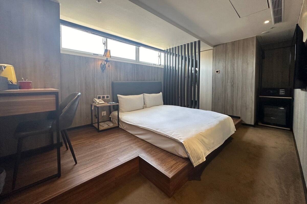 A modern hotel room with a large bed, a desk, and a chair. There is a window providing natural light and a view of the outside.