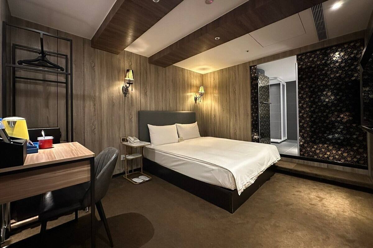 A modern hotel room with a large bed, a desk with a chair, a small table, and a walk-in shower with a floral tile backsplash.