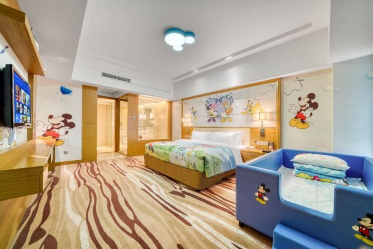 A hotel room with a bed, a desk, and a blue playpen with Mickey Mouse.