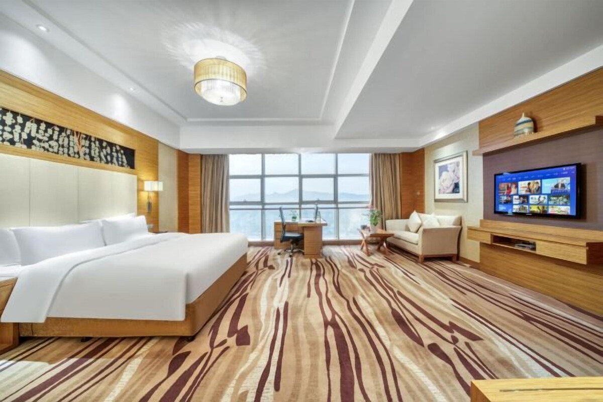 A hotel room with a large bed, a desk, a sofa, a TV, and a view of the mountains through the window.