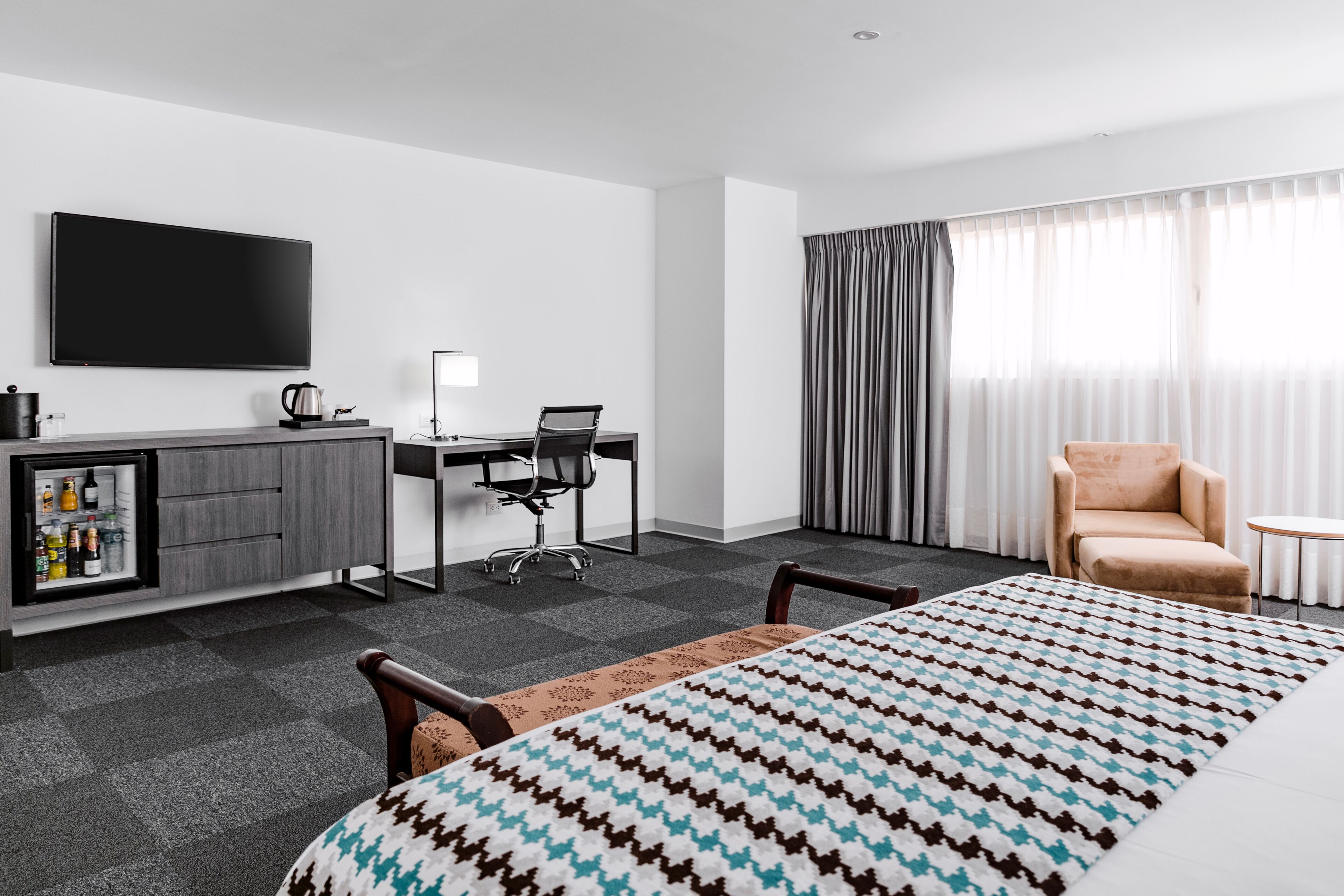 Habitación de hotel con cama, escritorio con silla, televisor, minibar y un amplio ventanal con cortinas.