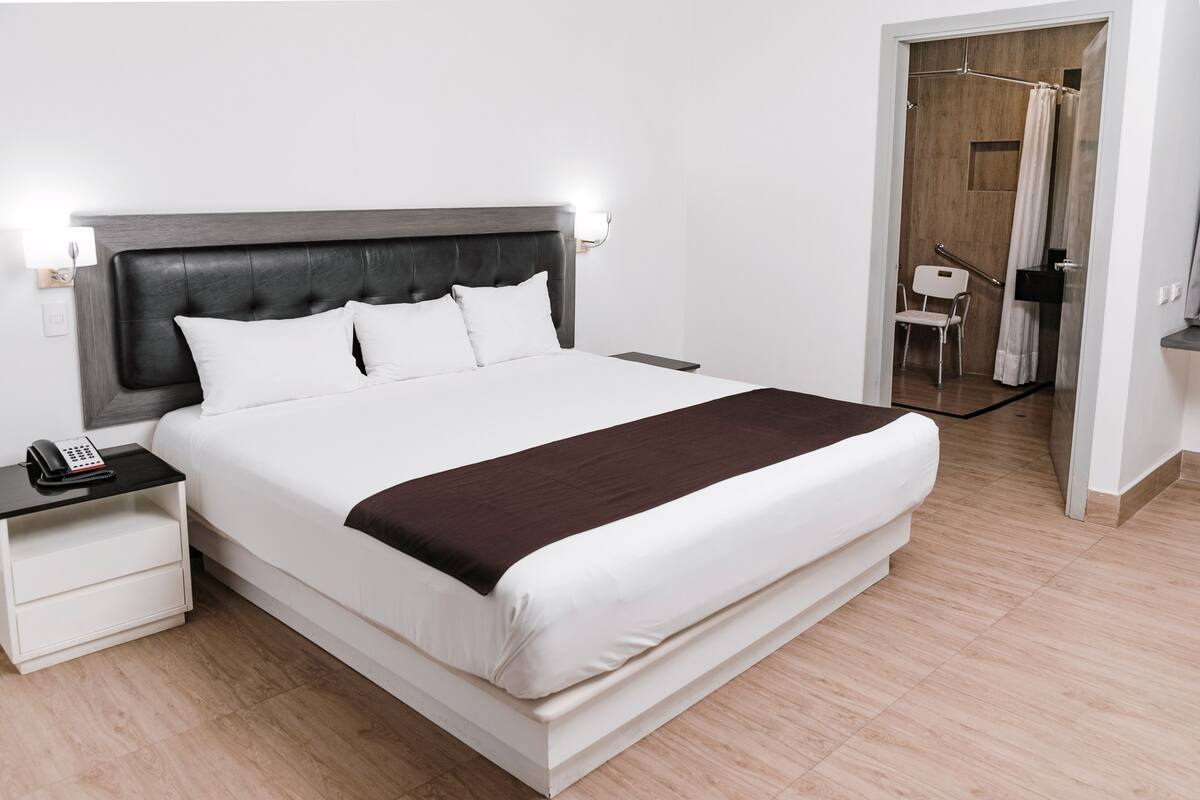Habitación de hotel moderna con una cama grande, mesitas de noche, un teléfono y una zona de ducha con asiento.