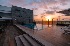 Un área de piscina en la azotea con vista al atardecer, un edificio moderno con un logotipo y mobiliario exterior.