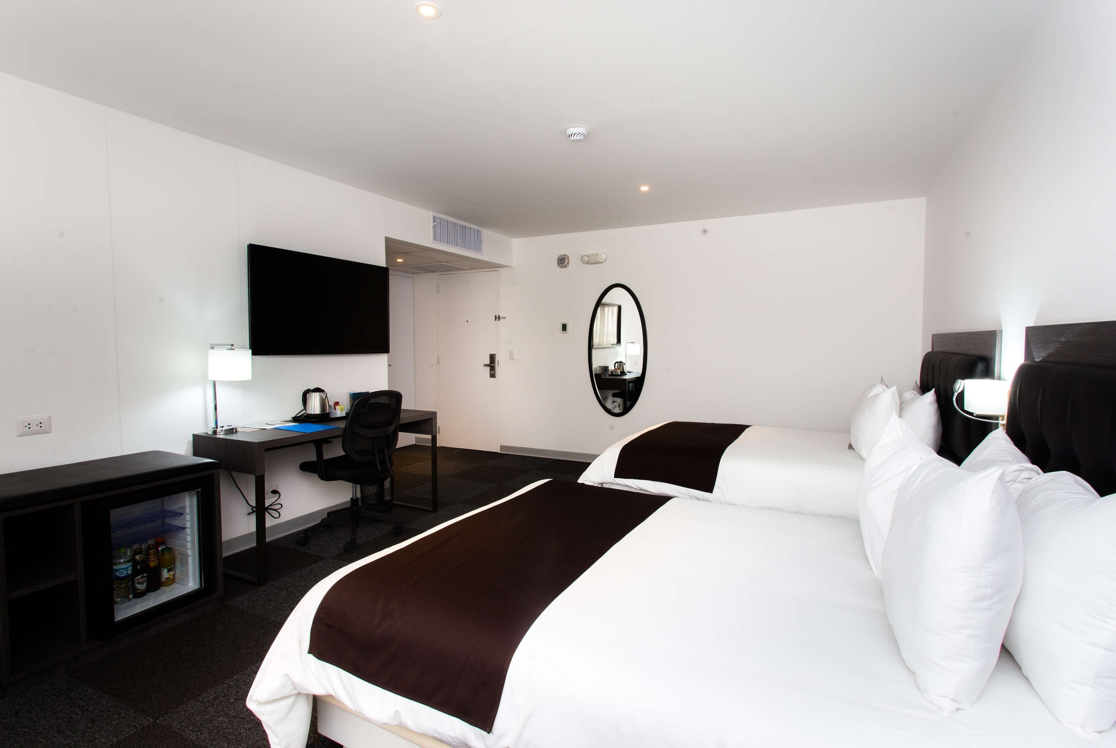 Habitación de hotel con cama, escritorio, silla, televisor, espejo y minibar.
