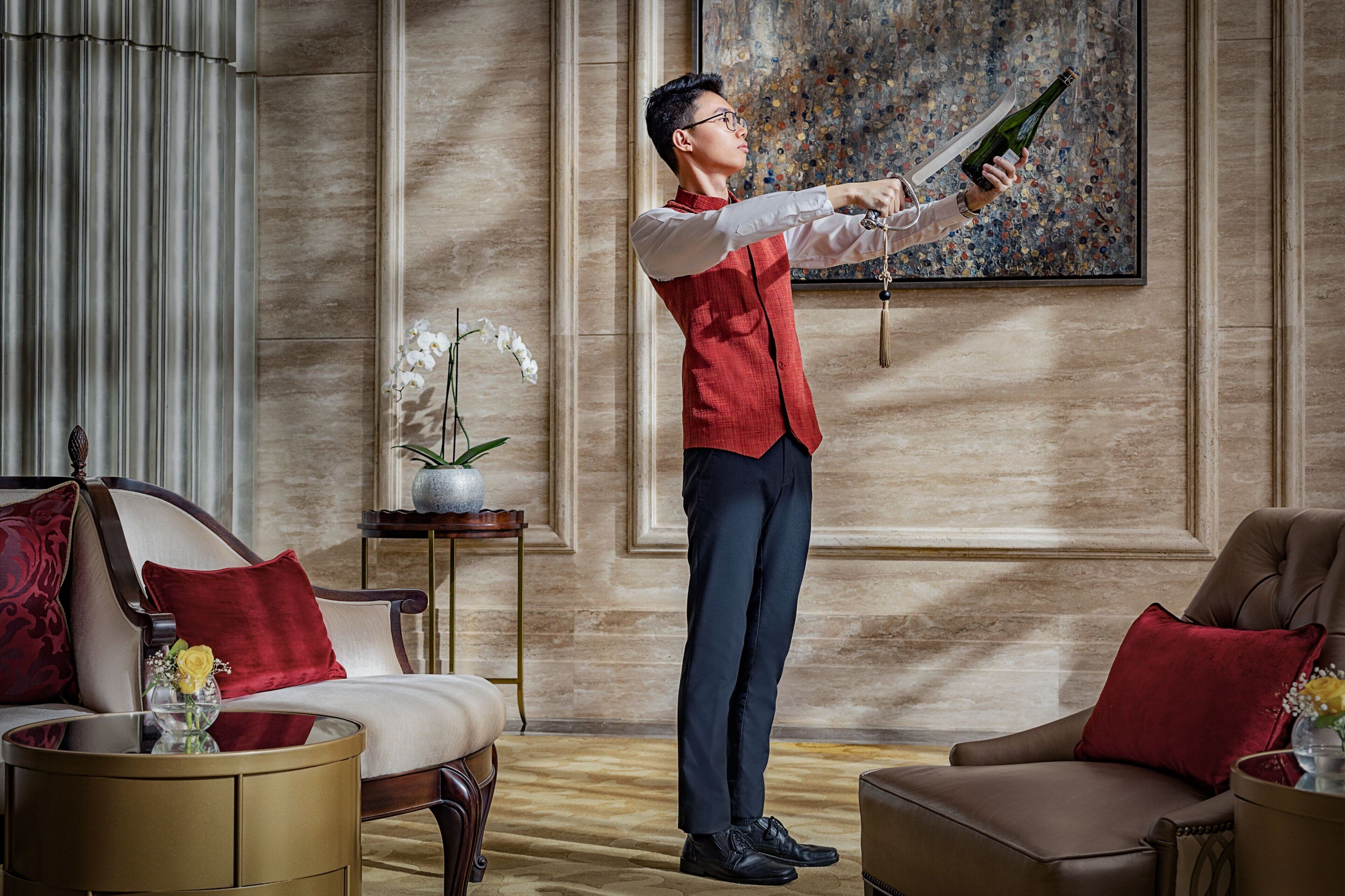 A man in a red vest and dark trousers holding a champagne bottle in a well-furnished room with a large painting on the wall, a potted plant, and a sofa with red cushions.